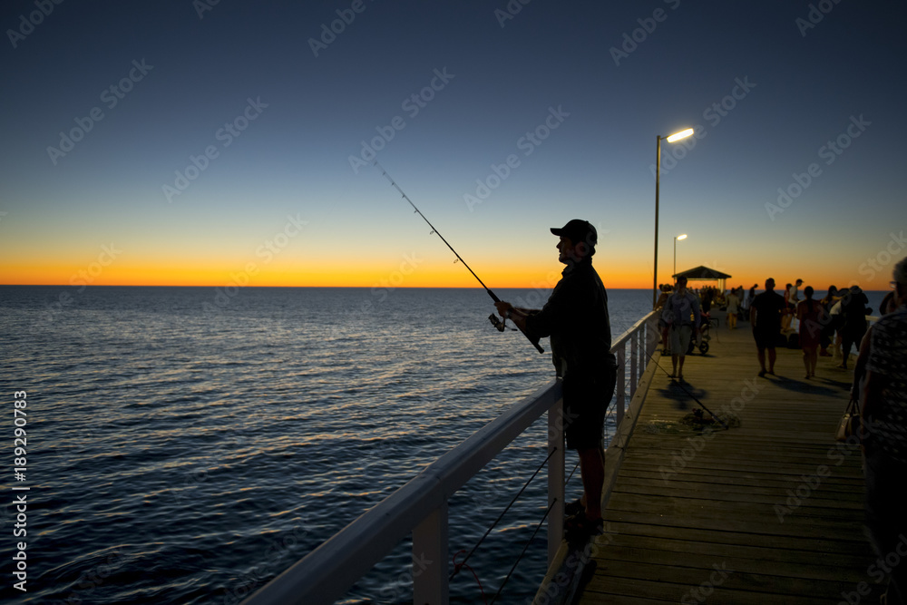 silhouette of fisherman with hat and fish rod standing on sea dock fishing at sunset with beautiful orange sky in vacations relax hobby