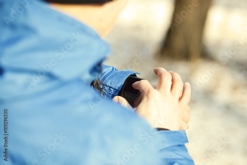 The young guy in the winter park in the sportswear is looking on his smartwatches