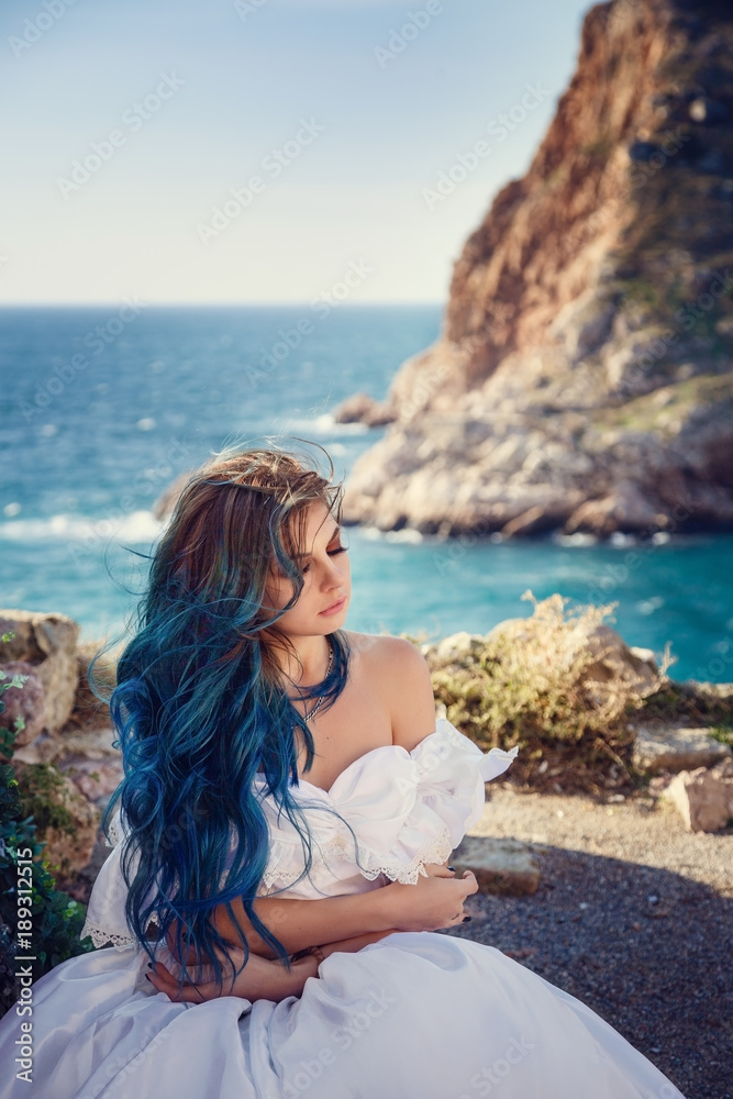 Young woman standing on cliff's edge and looking into a wide sea view. Wind Long Blue hair hairstyle.