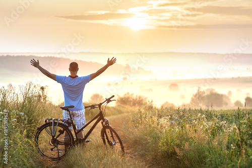 Young man standing near bicycle in morning sunrise with wonderful rays and morning mist during calm summer active day