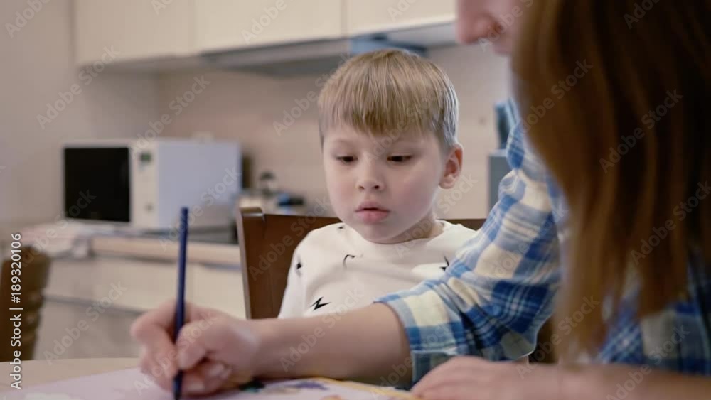 Close-up - Cute Little Curious Boy Looks At How His Beautiful Young Mother Draws And Asks Her About Something During Domestic Activities Close-up - Cute Little Curious Boy Looks At How His Beautiful Young Mother Draws And Asks Her About Something During Domestic Activities