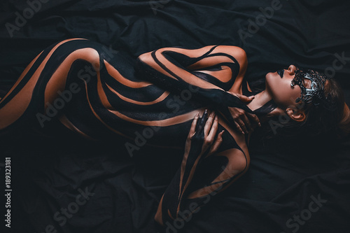 Beautiful girl with painted body and black mask in her face lies on bed. Body art.