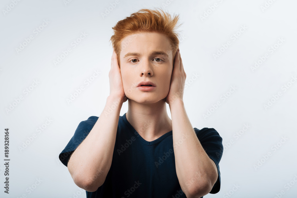 Loud noise. Appealing young tender guy placing hands on the ears while looking at the camera and standing on the light background Loud noise. Appealing young tender guy placing hands on the ears while looking at the camera and standing on the light background