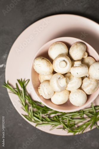 Diet, Italian Food Cooking, Vegetarian Concept. A bowl of fresh button mushrooms and a sprig of rosemary on a plate, dark concrete background, top view