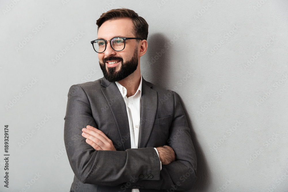 Close up portrait of pleased unshaved man in eyeglasses looking on camera with sincere smile, standing with arms folded isolated over gray background Close up portrait of pleased unshaved man in eyeglasses looking on camera with sincere smile, standing with arms folded isolated over gray background
