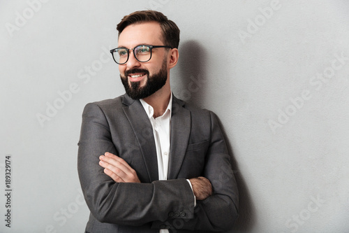 Close up portrait of pleased unshaved man in eyeglasses looking on camera with sincere smile, standing with arms folded isolated over gray background photo