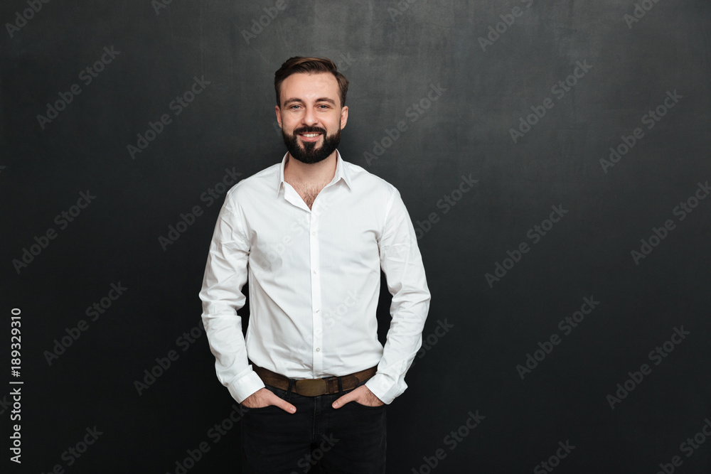 Portrait of young man in white shirt posing on camera with broad smile, and hands in pockets over dark gray background