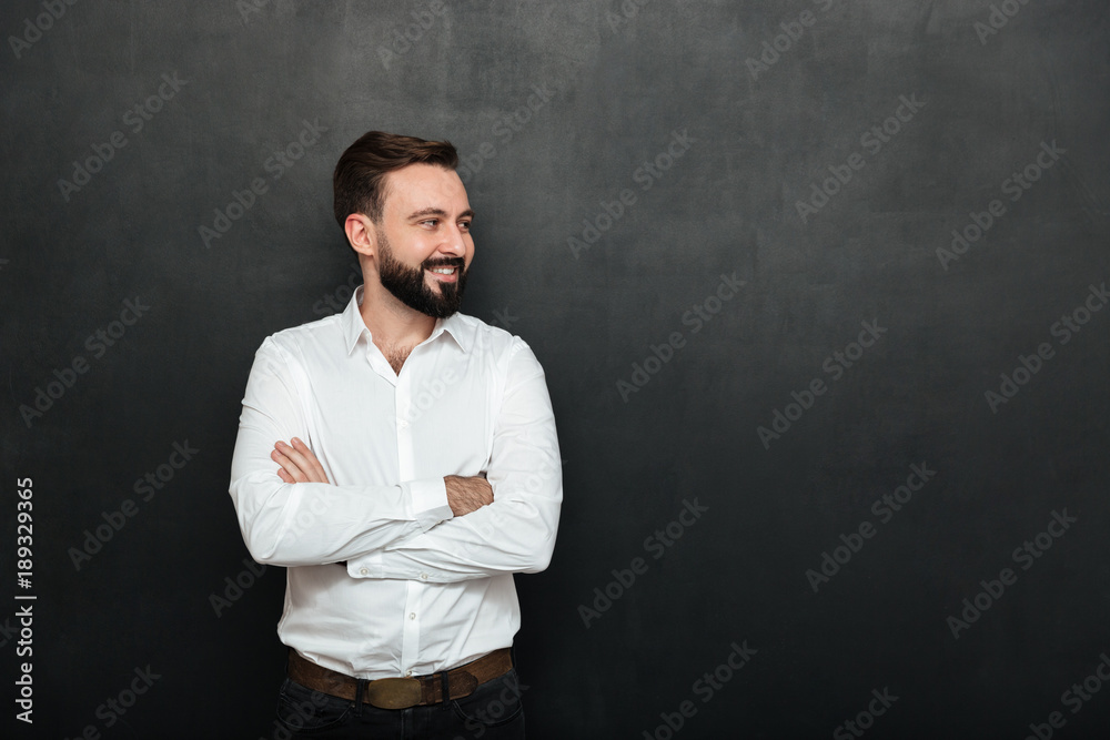 Portrait of positive man in white shirt standing with arms folded, and looking away over dark gray background copy space