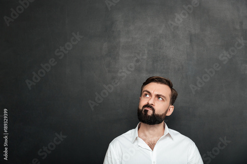 Portrait of brunette serious man in white shirt looking up with twisted face thinking or recalling, over dark gray background photo