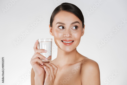 Beauty portrait of young happy woman with hair in bun drinking still water from transparent glass, isolated over white background