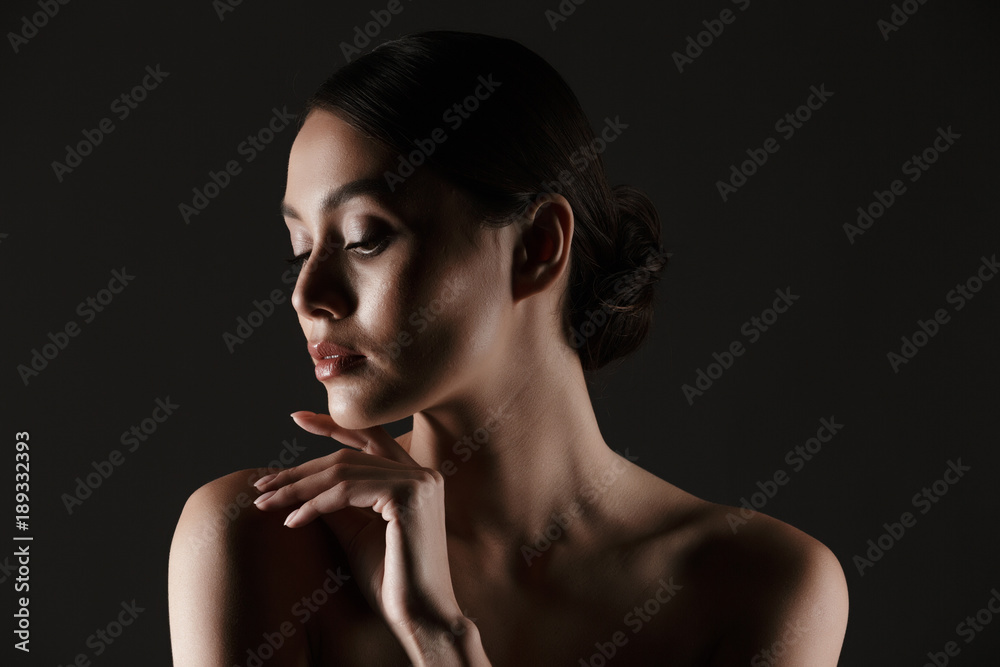 Naklejka premium Portrait of sensual beautiful woman looking aside while touching her chin in low lights, isolated over black background