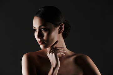 Portrait of feminine young woman with dark hair in bun posing at camera looking aside in low lights, isolated over black background
