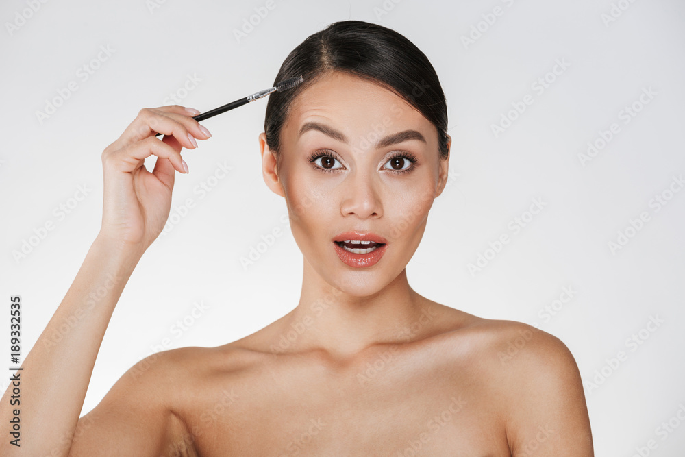 Beauty picture of gorgeous woman with hair in bun looking on camera and holding brush for eyebrows, isolated over white background