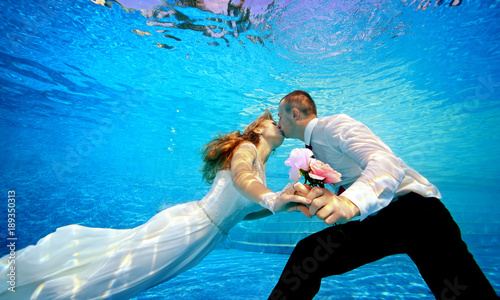 Lovers man and woman in wedding dresses kissing underwater in the pool and holding flowers in his hand. Horizontal orientation. A view from under the water from the bottom of the pool