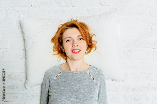 Red hair woman in gray shirt with a white pillow on the white brick wall. Work, home tiredness concept. photo
