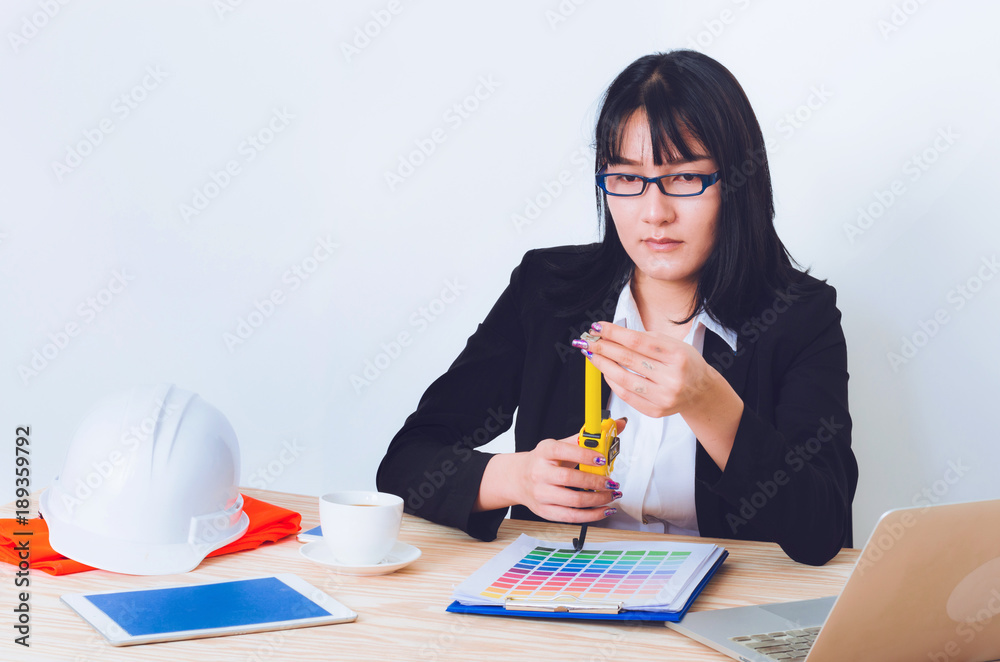 attractive business woman or engineer woman using measuring tape working on wood table in office with tablet, laptop, coffee cup, helmet and paper graph. engineering working design concept. attractive business woman or engineer woman using measuring tape working on wood table in office with tablet, laptop, coffee cup, helmet and paper graph. engineering working design concept.