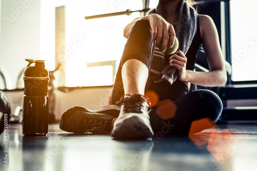 Sport woman sitting and resting after workout or exercise in fitness gym with protein shake or drinking water on floor. Relax concept. Strength training and Body build up theme. Warm and cool tone