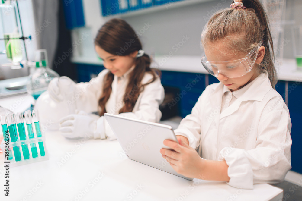 Two little kids in lab coat learning chemistry in school laboratory. Young scientists in protective glasses making experiment in lab or chemical cabinet. Working on a tablet. Two little kids in lab coat learning chemistry in school laboratory. Young scientists in protective glasses making experiment in lab or chemical cabinet. Working on a tablet.
