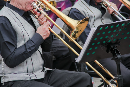 Old musican dressed in men's classic vest playing on a trombone in an orchestra, music stand behind him. Summer festival in a city