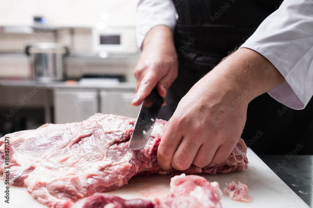 Restaurant chef in black apron cutting fresh lamb meat in the kitchen. Ingredient preparation before cooking a delicious meal