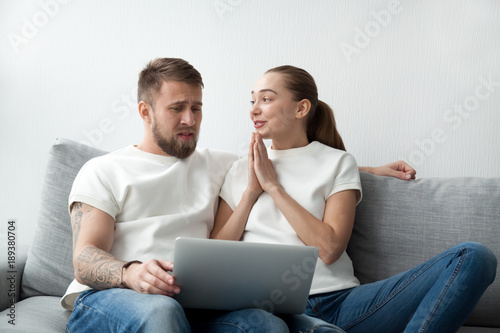Young couple doing online shopping with laptop sitting on sofa together, confused husband looking at screen while materialistic wife with begging hands asking husband please buy too expensive gift