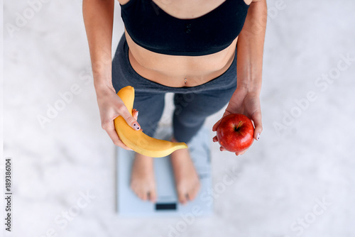 Sporty woman with a perfect body measuring body weight on electronic scales and holding a yellow banana and a red apple. Healthy low calorie meals,fruits. Diet, a healthy lifestyle. Weight loss.