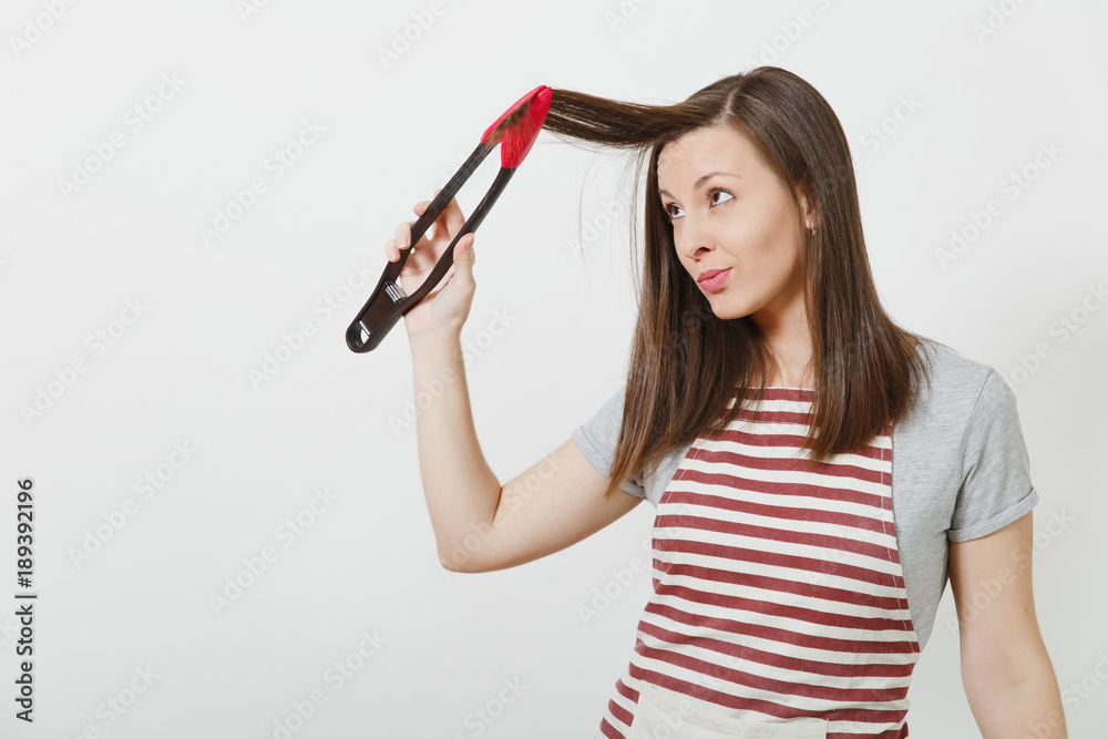 Young fun attractive smiling brunette caucasian housewife in striped apron isolated on white background. Beautiful housekeeper woman holding hair by kitchen tongs like styler curling hair. Young fun attractive smiling brunette caucasian housewife in striped apron isolated on white background. Beautiful housekeeper woman holding hair by kitchen tongs like styler curling hair.