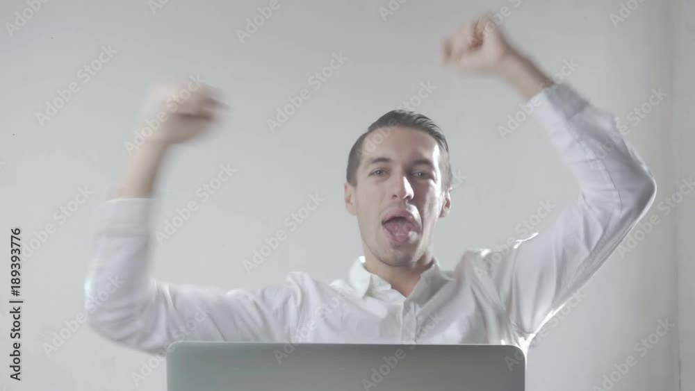 Young man in a white shirt very happy and rejoices sitting at laptop in office Young man in a white shirt very happy and rejoices sitting at laptop in office