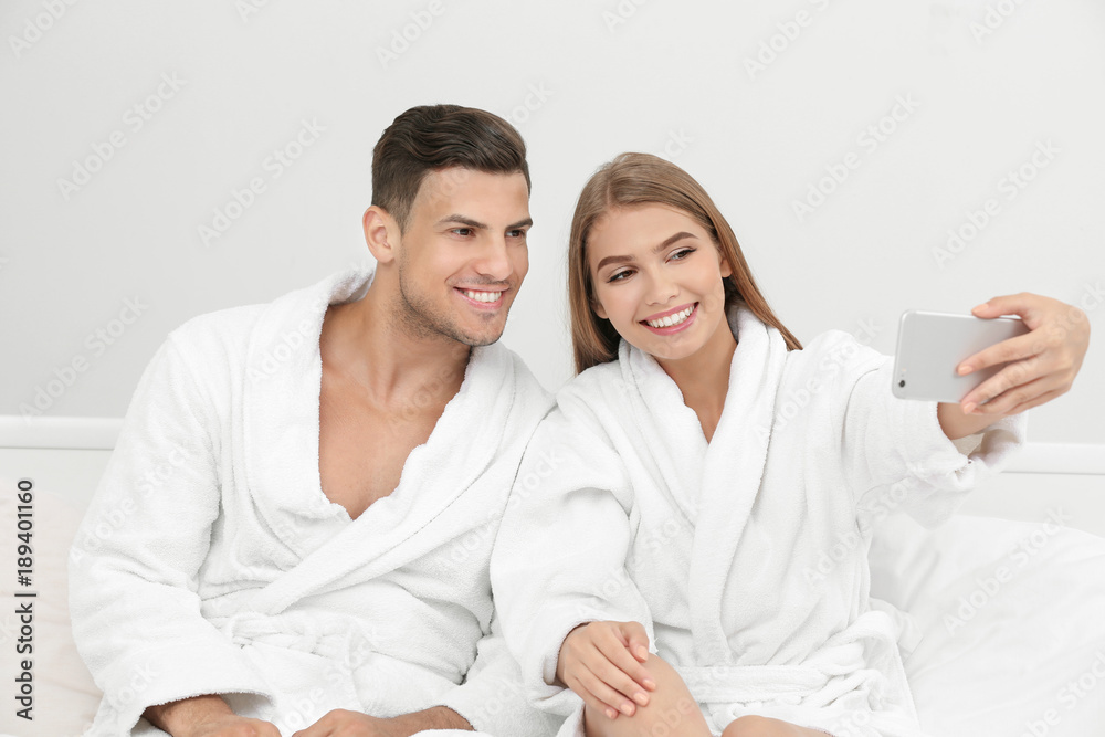 Young loving couple in bathrobes taking selfie on bed at hotel Young loving couple in bathrobes taking selfie on bed at hotel