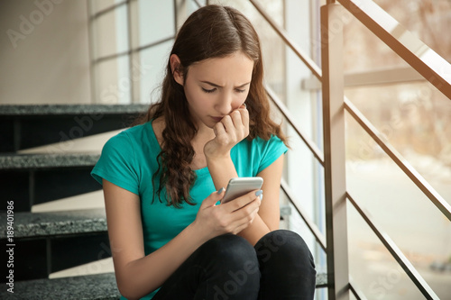 Upset teenage girl with smartphone indoors. Cyber bullying