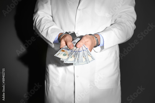 Male doctor in handcuffs holding bribe on black background