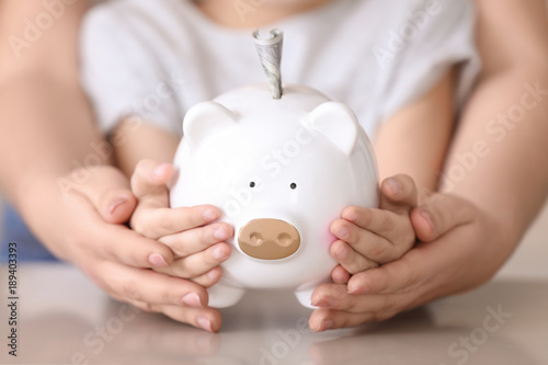 Little girl and young woman holding piggy bank with money at table