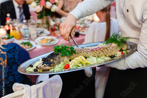 Dish of whole fish on a tray in the hand of the waiter. Male waiter handing a dish, and holding a tray with baked fish and garnish, close-up