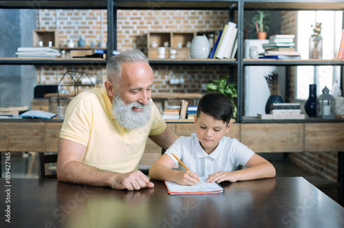 He is so smart. Waist up shot of a joyful senior gentleman grinning broadly while sitting at a table and doing a home assignment with his grandson.