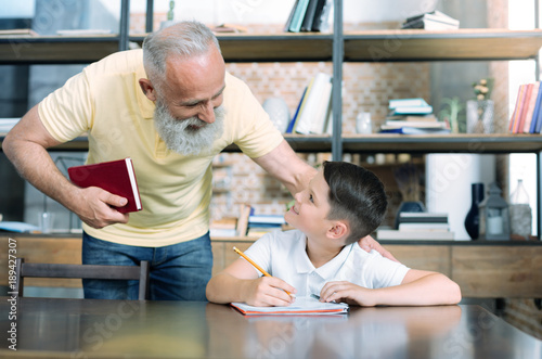 Do you need some help. Thoughtful elderly gentleman smiling while addressing to his smart little grandchild and helping him with homework. © zinkevych