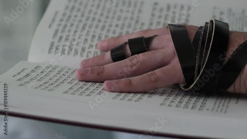 Hand of young jewish man with a tefillin on his arm, holding a psalms book, while reading a pray at a jewish ritual.