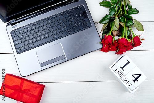 Top view of bouquet of red roses, red gift box, wood calendar february 14 and open laptop computer on white table. Greeting card for Valentines Day. Flat lay © mirage_studio