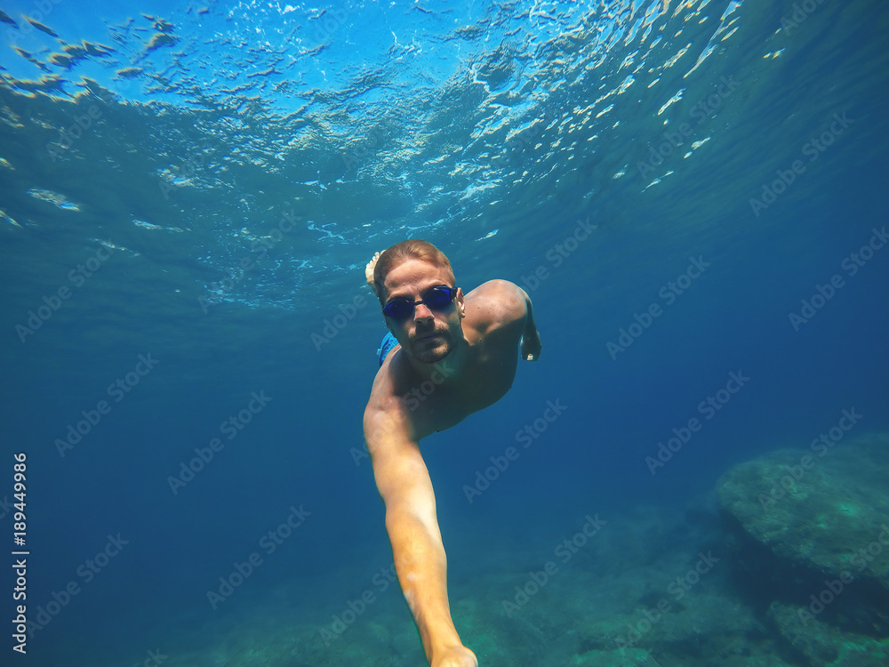 Underwater photo of a young sportive active man diving in the turquoise exotic sea near the rocks and taking a selfie with a stick for summer holidays.