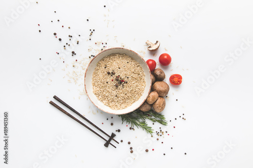 flat lay composition of raw rice in bowl with mushrooms and tomatoes on white surface with chopsticks