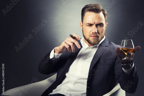 Handsome young bearded man with  brandy glass with cognac and cigar. Perfect hair cut. Wearing suit and white shirt