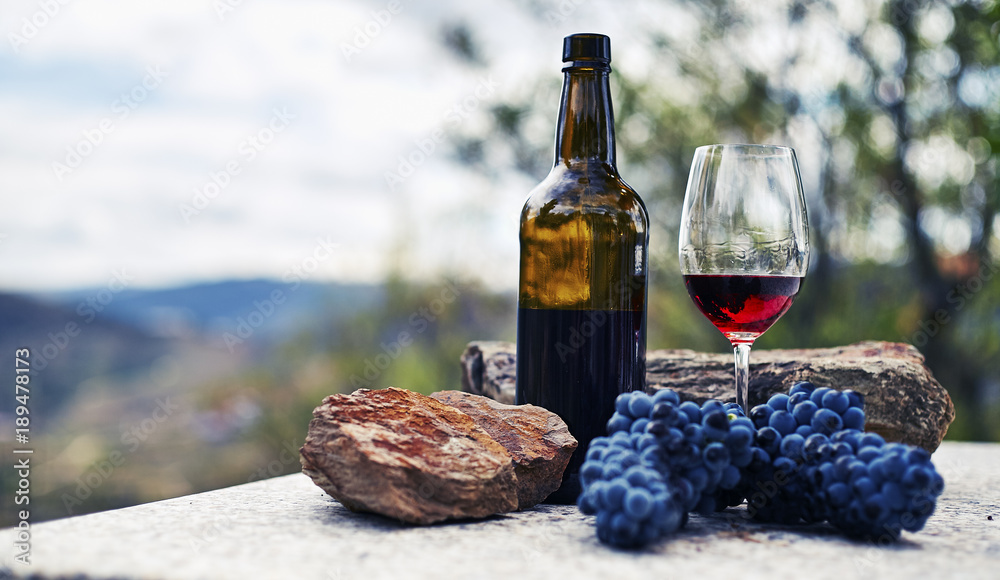 Glass bottle and glass of red wine on stone table with bunch of grapes and majestine background of mountains Glass bottle and glass of red wine on stone table with bunch of grapes and majestine background of mountains