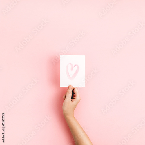 Valentine's Day composition. Female hand holding card with heart symbol on pale pink background. Flat lay, top view Love concept.