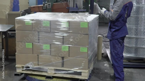 Branch of boxes are being packed with polyfirm on the plant. The block is turning on the special machine and every continer with production has green sticker.Male worker in blue uniform is standing photo