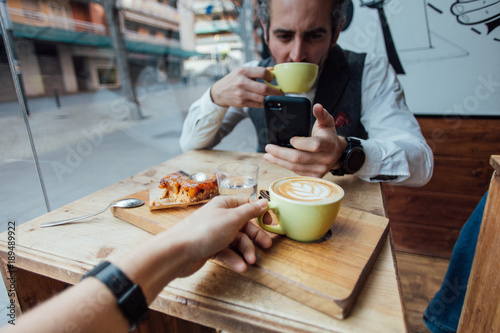 Happy and loving hipster millennial couple, shares lunch or brunch at morning trendy cafe or coffeeshop, drink latte or espresso, eat sweet dessert, easy going lifestyle of freelancers or bloggers