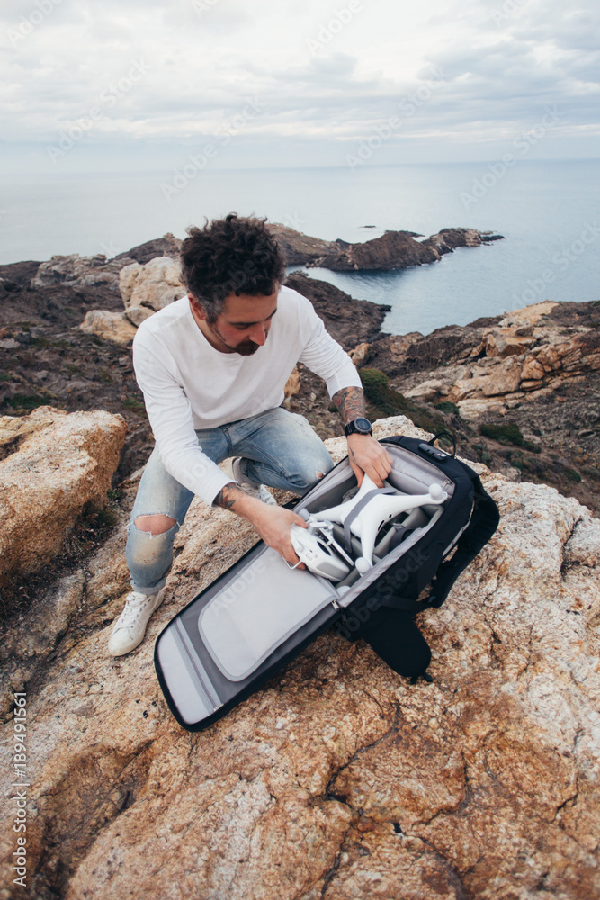 Fototapeta premium Professional stock aerial photographer, urban adventurer and location explorer, traveler unpacks backpack with expensive equipment and drone, ready to fly over oceanside and inspiring scenery