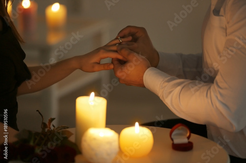 Man putting engagement ring on fiancee's finger while sitting at table with burning candles