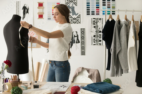 Woman fashion designer measuring mannequin, seamstress holding tape working with dummy in cozy creative design studio or tailor shop, dressmaking and sewing concept, exclusive unique clothes creation