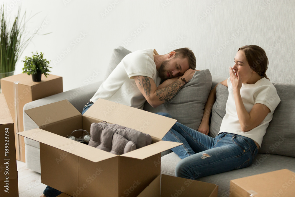 Young tired couple feeling fatigue on long hard moving out day, exhausted restless man having break in new home while sleepy woman yawning on sofa after packing staff in boxes preparing to relocate Young tired couple feeling fatigue on long hard moving out day, exhausted restless man having break in new home while sleepy woman yawning on sofa after packing staff in boxes preparing to relocate