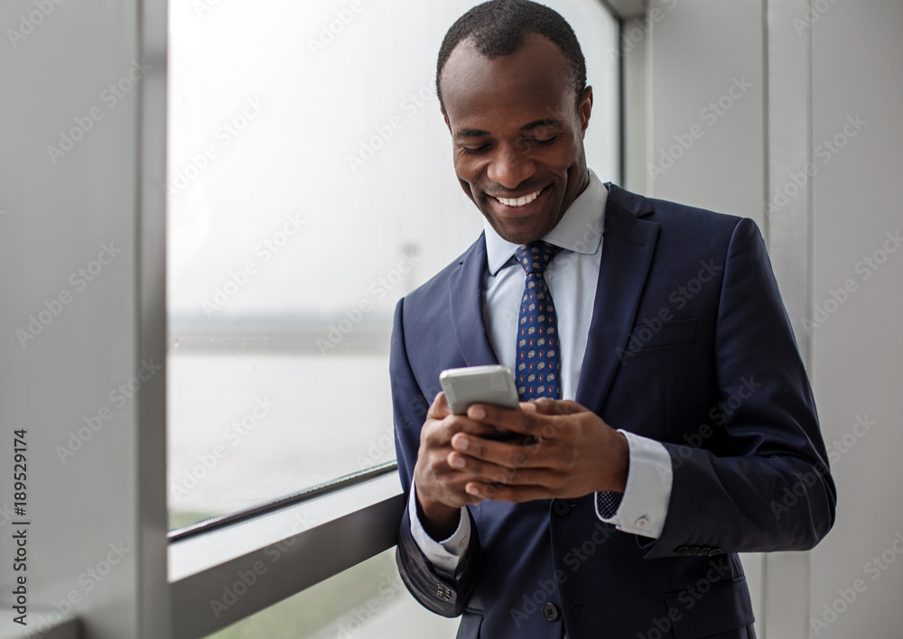 Chatting with pleasure. Portrait of happy delighted young entrepreneur is standing in light office against window and holding mobile phone. He is reading messages on gadget with smile. Copy space Chatting with pleasure. Portrait of happy delighted young entrepreneur is standing in light office against window and holding mobile phone. He is reading messages on gadget with smile. Copy space