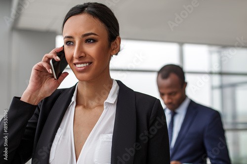 Successful negotiations. Portrait of gorgeous elegant businesswoman is standing and talking on smartphone. She is looking aside with smile while african man is standing in background